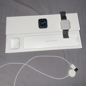 Apple Watch med silver mesh-armband - Säljer en Apple Watch med fyrkantig display och ett snyggt silverfärgat mesh-armband i metall. Klockan har digital touchskärm och kommer med originalförpackning samt laddkabel. Perfekt för dig som gillar tech och vill ha koll på allt direkt på handleden.