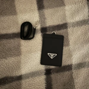 Svart plånbok med rem - Svart plånbok med avtagbar rem och metalllogo i silver på framsidan. Plånboken har en stilren, rektangulär form och fack med transparent plast på baksidan för kort eller ID. Materialet är skinn med en snygg textur. Pris går att deskuteras via snabb affär