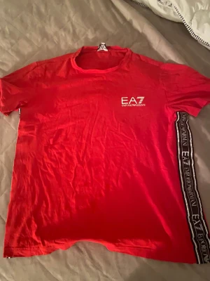 Röd EA7 Emporio Armani t-shirt - Snygg röd t-shirt från EA7 Emporio Armani med vit logga på bröstet och svart/vit logoband längs sidan. Klassisk passform och rund hals. Tillverkad i mjuk bomull som känns skön mot huden. Perfekt för dig som gillar streetwear och vill sticka ut.