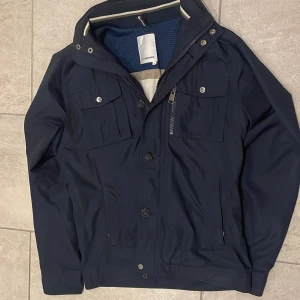 Mörkblå vindjacka från J.Lindeberg - Snygg mörkblå vindjacka från J.Lindeberg med praktiska fickor, dragkedja och knappar framtill. Jackan har hög krage, justerbara detaljer i midjan och meshfoder på insidan. Perfekt för dig som gillar stilren och funktionell design. Är  i storlek m men sitter mer som s