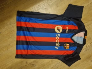 FC Barcelona Lewandowski fotbollströja - Säljer en FC Barcelona matchtröja med Lewandowski och nummer 9 på ryggen. Tröjan har breda blå och röda ränder, guldiga tryck och klubbmärke på bröstet. Spotify-logga på framsidan och UNHCR-tryck på ryggen. Tillverkad i lätt polyester, perfekt för träning eller match.