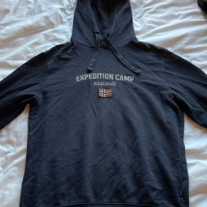 Svart hoodie från Napapijri Expedition - Svart hoodie från Napapijri Expedition Camp med tryck och norsk flagga på bröstet. Tröjan har huva med snörning och långa ärmar. Mjuk bomullskvalitet och ribbade muddar. Perfekt för dig som gillar outdoor-vibe och streetstyle.