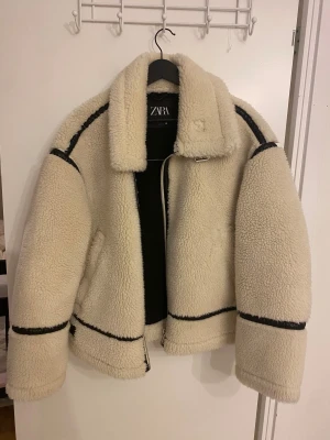 Teddy jacka Zara  - Säljer en beige teddyjacka från Zara med svarta skinnimitation-detaljer och grov krage. Jackan har dragkedja, bälte vid halsen och stora fickor. Insidan är svart och jackan har en loose passform, perfekt för kalla dagar.