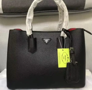 Svart handväska från Prada i skinn - Snygg svart handväska från Prada med stilren design och röd insida. Väskan är tillverkad i strukturerat skinn och har två rejäla handtag, samt en praktisk dragkedja upptill. Perfekt för dig som vill ha en lyxig och klassisk accessoar med plats för allt du behöver.