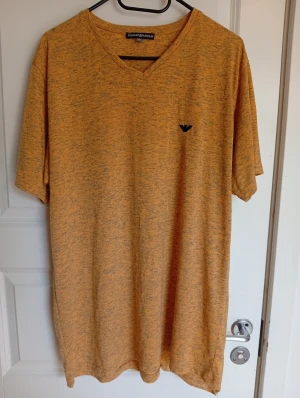 Orange melerad t-shirt - Snygg orange melerad t-shirt från  med v-ringad hals och korta ärmar. T-shirten har ett svart broderat logomärke på bröstet och är gjord i mjuk bomull. Perfekt för dig som gillar stilrena och bekväma plagg med lite extra attityd. Liten i storlek