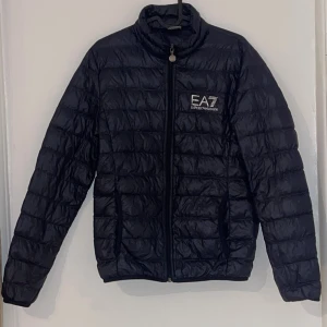 Mörkblå EA7 Emporio Armani pufferjacka - Snygg mörkblå pufferjacka från EA7 Emporio Armani med quiltad design och hög krage. Jackan har dragkedja framtill, två sidofickor och tydligt EA7-logga på bröstet. Perfekt för dig som vill ha en stilren och modern look under kyliga dagar.