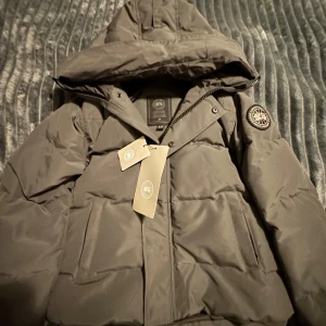 Mörkgrå pufferjacka från Canada Goose - Säljer en mörkgrå pufferjacka från Canada Goose med huva och snygg patch på ärmen. Jackan har dragkedja och knappar framtill, två stora fickor och quiltad design. Perfekt för kalla dagar och har en clean, modern look.Model McMillan 