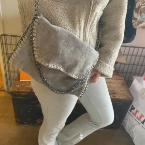 Grå axelväska med kedjedetaljer - Snygg grå axelväska i mocka med grova kedjedetaljer längs kanterna och som axelrem. Väskan har en mjuk, slouchy form och dekorativa sömmar runt locket. Perfekt för dig som gillar stilrena accessoarer med lite edge.