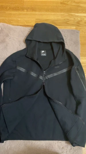 Svart vindjacka från Nike med huva - Svart Nike vindjacka med huva och hel dragkedja framtill. Jackan har diskreta grå detaljer i form av ränder över bröstet och på ärmen, samt en extra ficka med dragkedja på vänster arm. Perfekt för dig som gillar sportig och clean stil.