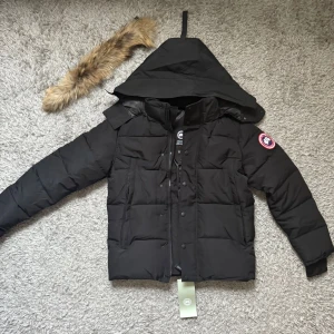 Canada Goose jacka svart - Säljer en svart dunjacka från Canada Goose med avtagbar huva och pälskant. Jackan har klassisk patch på ärmen, dragkedja och knappar framtill samt ribbade muddar. Perfekt för kalla vinterdagar och riktigt snygg streetstil.