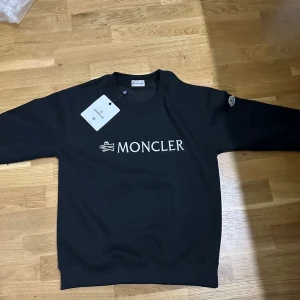 Svart sweatshirt från Moncler - Svart sweatshirt från Moncler med vit broderad logga på bröstet och Moncler-märke på ärmen. Tröjan har rund hals, långa ärmar och ribbade muddar. Perfekt för dig som gillar stilrena och exklusiva plagg med tydlig branding.