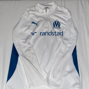 Vit långärmad Marseille Puma tröja - Säljer en vit långärmad fotbollströja från Puma med Olympique de Marseille-logga och blå detaljer på sidorna. Tröjan har halv dragkedja fram, tryckt Puma-logga och sponsortryck. Materialet är lätt och ventilerande polyester, perfekt för träning.
