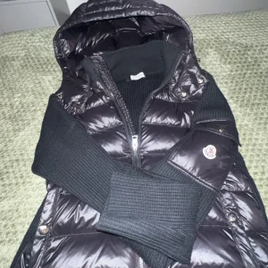 Svart Moncler pufferjacka med huva - Säljer en svart cardigan från Moncler med stickade ärmar och detaljer. Jackan har en stor, vadderad huva och dragkedja framtill. Moncler-logga på ärmen och snygga ribbade muddar. Perfekt för dig som vill ha en cool och varm jacka med streetkänsla. Är 175cm och väger 67 kg och passar bra/