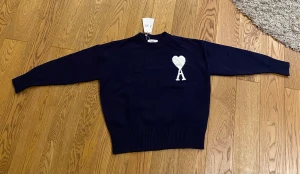 Ami Paris mörkblå - Snygg mörkblå stickad tröja från Ami med ett vitt hjärta och ett stort A broderat på bröstet. Tröjan har rund hals, ribbade muddar och lång ärm. Perfekt för dig som gillar stilrena detaljer och vill ha något som sticker ut lite extra.