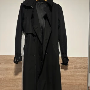Svart trenchcoat från Zara Woman - Snygg svart trenchcoat från Zara Woman med klassisk dubbelknäppning, bälte i midjan och detaljer vid ärmsluten. Jackan har lång passform och är perfekt för dig som gillar stilrena och tidlösa plagg. Materialet är mjukt och känns lyxigt.