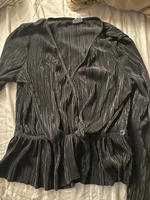 Svart omlottopp från Gina Tricot - Säljer en svart omlottopp från Gina Tricot i storlek S. Toppen har ett snyggt plisserat tyg med glansig finish, volang vid midjan och långa ärmar. Perfekt för dig som gillar trendiga detaljer och vill ha en cool look.
