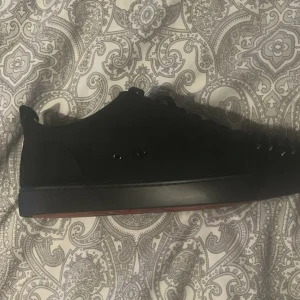 Svarta Christian Louboutin sneakers med nitar - Säljer ett par svarta sneakers från Christian Louboutin med coola svarta nitar på tån. Skorna har snörning och en platt sula med den klassiska röda undersidan. Materialet är mocka och designen är stilren men edgy, perfekt för dig som vill sticka ut.