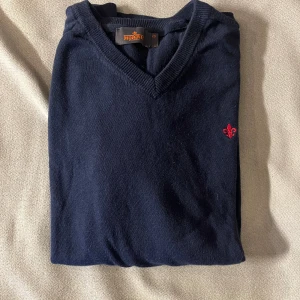 Morris Marinblå v-ringad tröja - Snygg marinblå v-ringad tröja från Morris med röd broderad logga på bröstet. Tröjan är långärmad och har ribbade muddar vid ärmslut och nederkant. Perfekt för dig som gillar klassisk stil med en modern touch.