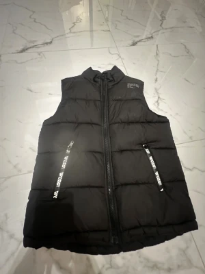 Svart teckväst från Kappahl/woxo - Svart väst från  Kappahl med hög krage och två fickor med dragkedja där det står WOXO på . Västen har quiltad design och stängs med en rejäl dragkedja framtill. Perfekt för lager-på-lager under kyliga dagar.