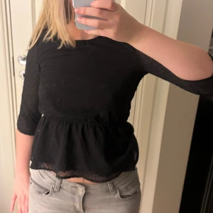 Svart blus med prickigt mesh - Säljer en svart blus med diskret prickigt mesh-tyg och volang nertill. Toppen har trekvartsärm och en lätt transparent känsla på ärmarna. Sitter snyggt i midjan och ger en feminin siluett. Perfekt att styla med jeans eller kjol.