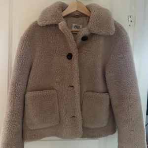 Beige teddyjacka från Zara, S - Säljer en beige teddyjacka från Zara i storlek S. Jackan har fluffigt material, stora framfickor och svarta knappar framtill. Den har en bred krage och är långärmad, perfekt för kalla dagar. Jackan har en loose passform och är supermysig att bära.
