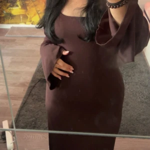 Brun långklänning med vida ärmar som framhäver midjan med knutarna på baksidan - Säljer en elegant brun långklänning med snygg, figurnära passform och vida ärmar. Klänningen har en enkel, stilren design med rundad halsringning och mjukt, stretchigt material som sitter skönt mot kroppen. Perfekt för dig som gillar minimalistisk och chic stil.