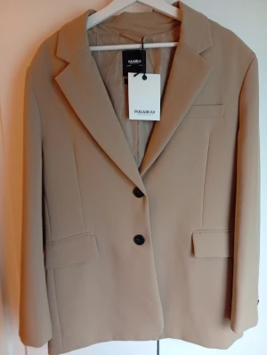 Beige kavaj från Pull&Bear M - Stilren beige kavaj från Pull&Bear i storlek M. Oversize modell Klassisk modell med två svarta knappar framtill, fickor med lock och snygg krage. Tillverkad av återvunnen polyester, perfekt för dig som gillar minimalistisk och modern stil. Extra knapp medföljer.