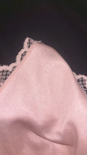 Rosa spetsig linnetopp med rosett - Söt ljusrosa linnetopp med tunna axelband, spetsdetaljer och liten rosett framtill. Toppen har glansigt tyg med diskreta ränder och spets över bysten och sidorna. Perfekt för en romantisk look eller som layering-piece. Finns små fläckar inuti och lite nopprig.