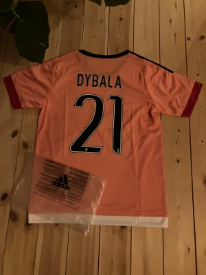 Dybala #21 Juventus tröja - Bilderna visar en rosa Juventus fotbollströja från Adidas med Dybala och nummer 21 på ryggen, samt Jeep-logga på framsidan. Ingen sko finns med på bilderna. Inte riktig men jättebra kvalite