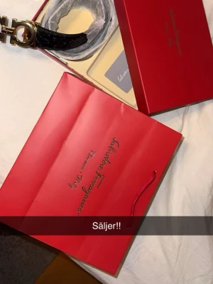 Svart bälte  från Salvatore Ferragamo - Säljer ett svart skärp från Salvatore Ferragamo med elegant guldfärgat spänne. Skärpet kommer i originalförpackning med röd presentpåse och ask. Perfekt accessoar för att lyfta din outfit.