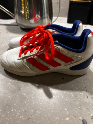 Adidas fotbollsskor barn  (inomhus sko) SALA - Säljer ett par Adidas fotbollsskor med vit bas, röda ränder och snörning samt blå detaljer vid häl och krage. Skorna har platt sula och är märkta med 'SALA' baktill. Materialet är en mix av skinn och syntet, perfekt för inomhusspel.