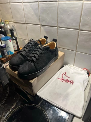 Christian Louboutin Jr Flat  - Snygga svarta sneakers från Christian Louboutin med klassisk röd sula och svart snörning. Utsidan är i mocka och insidan har en beige skinnfodring. Skorna har rund tå och platt sula, perfekt för dig som gillar exklusiva märken och stilren design. Kan bytas mot lanvins eller andra märkesskor
