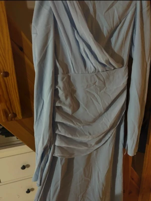 Ljusblå långklänning med snörning - Säljer en elegant ljusblå långklänning med snygga draperingar och omlott-detaljer framtill. Klänningen har långa ärmar och en cool snörning i ryggen som man kan göra de tajtare med och  ger en unik look. Materialet känns mjukt och lätt, perfekt för dig som vill sticka ut.