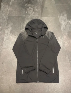 Svart vindjacka från Under Armour - Svart vindjacka från Under Armour med huva och hel dragkedja framtill. Jackan har diskreta mönstrade paneler på axlarna och reflekterande detaljer vid fickorna. Tillverkad i lätt polyester, loose fit och perfekt för utomhusaktiviteter.