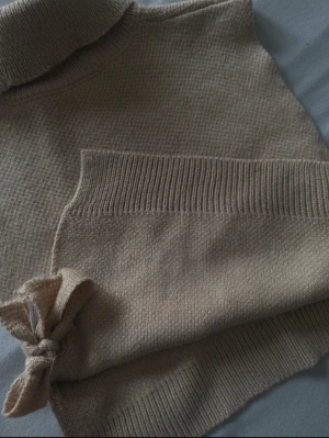 Beige stickad polotröja med knyt - Säljer en beige stickad polotröja utan ärmar med snygga knytband på sidorna. Tröjan har en hög polokrage och ribbade detaljer vid nederkant och hals. Perfekt att styla lager på lager för en chill och trendig look. Funkar att ha den på skjortor, vanliga tröjor eller bara så den är jätte skön!