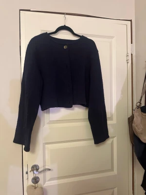Mörkblå croppad kofta med knapp - Säljer en mörkblå croppad kofta med lång ärm och rund hals. Koftan har en enkel design med knappar framtill och är tillverkad i ett mjukt stickat material. Perfekt att slänga över en t-shirt eller linne för en chill look.