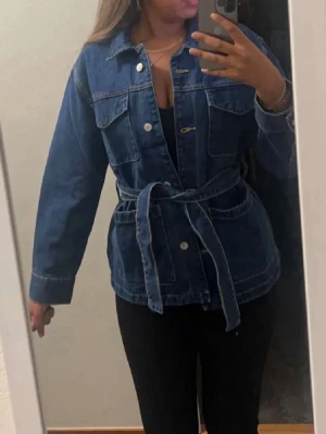 Blå jeansjacka med bälte - Säljer en snygg blå jeansjacka med bälte i midjan och knappar framtill. Jackan har långa ärmar, två bröstfickor med lock och en klassisk krage. Perfekt för dig som vill ha en chill och trendig look.