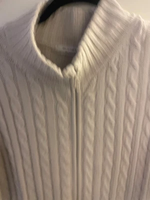 Beige stickad kofta från Sand - Säljer en beige stickad kofta från Sand med snygg kabelstickad design och ribbade muddar. Koftan har hel dragkedja framtill och en liten metallbricka vid nederkanten. Perfekt för lager-på-lager under kyliga dagar.