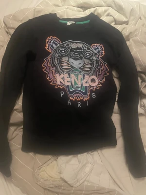Svart Kenzo sweatshirt med tiger - Svart sweatshirt från Kenzo i storlek XS med ikonisk broderad tiger och färgglada detaljer på bröstet. Tröjan har rund hals, långa ärmar och ribbade muddar. Perfekt för dig som gillar streetwear och vill sticka ut med ett coolt motiv.