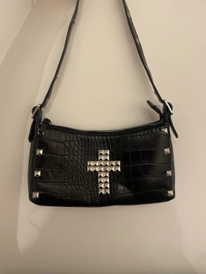 Svart axelväska med nitar - Cool svart axelväska i croco-mönstrat skinnimitation med silvriga nitar formade som ett kors på framsidan. Väskan har justerbar rem med metallhål och dragkedja upptill. Perfekt för dig som gillar edgy detaljer och unik stil.