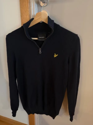 Mörkblå stickad tröja Lyle & Scott - Snygg mörkblå stickad tröja från Lyle & Scott med halv dragkedja och gul broderad logga på bröstet. Liten i storlek så mer som XS