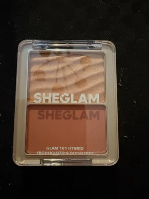 SHEGLAM Highlighter & Rouge Duo - Säljer en SHEGLAM Glam 101 Hybrid duo med highlighter och rouge. Highlightern har en skimrande champagnefärg med vågig yta, och rougen är matt i en varm persikoton. Kommer i en smidig plastförpackning med genomskinligt lock.