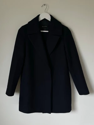 Mörkblå kappa från Massimo Dutti - Stilren mörkblå kappa från Massimo Dutti i klassisk rak modell med bred krage och långa ärmar. Kappan är tillverkad i ullblandning och har en slät yta utan synliga knappar eller detaljer. Perfekt för dig som gillar minimalistisk och tidlös stil. 5L1