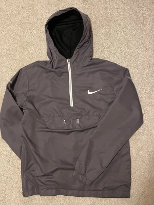 Nike Air Vindjacka - Nike Air vindjacka i grått med vit dragkedja och huva. Jackan har reflekterande detaljer på ärmarna, tryckta loggor på ryggen och en stor ficka framtill med AIR-tryck. Perfekt för dig som gillar sportig stil och vill ha något lätt och snyggt. Den har ett eller två små slitage på ena armen, knappt synliga. Passar också S.