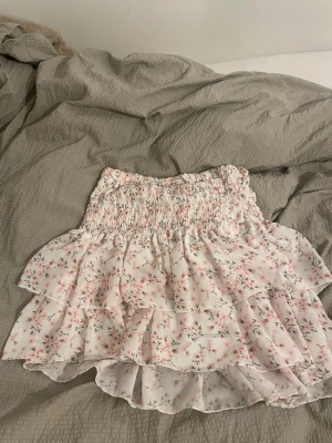 Blommig volangkjol i ljusrosa - Supersöt kort kjol med volanger och blommigt mönster i ljusrosa och grönt. Kjolen har smock i midjan för skön passform och är gjord i ett lätt, luftigt material som passar perfekt till varma dagar. Perfekt för dig som gillar romantisk och feminin stil.