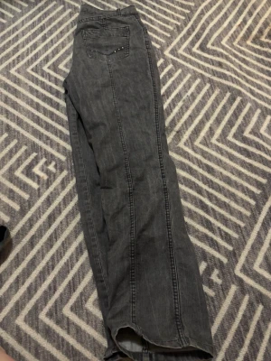 Grå bootcut jeans med nitar - Säljer ett par grå jeans med bootcut-snitt och snygga nitar på bakfickan. Jeansen har klassisk femficksdesign, knapp och dragkedja framtill samt bälteshällor. Materialet är jeans och färgen är mörkgrå, vilket ger en cool och avslappnad vibe. Storlek står ej men passar mig som S.