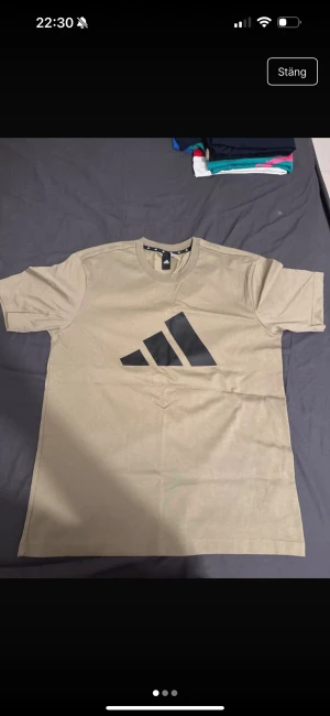 Beige t-shirt från Adidas med logga - Säljer en beige t-shirt från Adidas i storlek M. T-shirten har en stor svart Adidas-logga på framsidan, rund hals och klassisk passform. Materialet är mjuk bomull och modellen är kortärmad, perfekt för chill dagar eller sport.