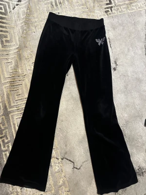 Svarta mjukisbyxor med strassfjäril :) - Svarta mjukisbyxor i sammet med bred resår i midjan och en snygg fjäril i strass på höften. Byxorna har bootcut-snitt och känns riktigt mjuka och sköna. Perfekt för dig som gillar chill vibe och lite extra bling men ett litet slitage i bakdelen som knappt syns :)