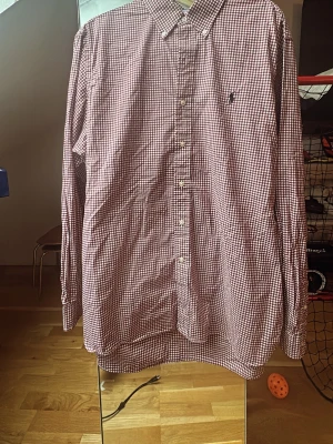 Rutig skjorta från Ralph Lauren - Säljer en snygg röd och vit rutig skjorta från Ralph Lauren i storlek L. Skjortan är tillverkad i 100% bomull med stretch och har klassisk krage, knappar framtill och lång ärm. Perfekt för dig som gillar stilrena och tidlösa plagg.