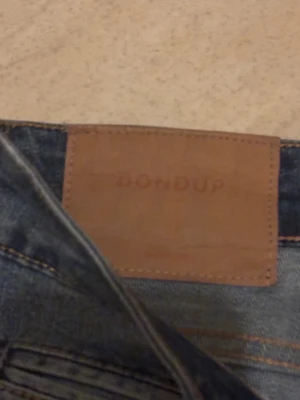Blå jeans från Dondup George  - Säljer ett par blå jeans från Dondup med snygga slitningar och distressed detaljer. Modellen har fem fickor, smal passform och klassisk knappgylf. Jeansen har en mörk tvätt med ljusare partier och är tillverkade i mjukt denimtyg.